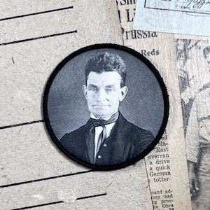 May include: Black and white photo of a man wearing a suit and tie. The photo is printed on a round, black fabric patch.