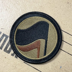 Könnte beinhalten: Ein gestickter schwarzer und brauner Aufnäher mit einem stilisierten Flaggenmotiv. Die Flagge ist schwarz mit einem braunen Griff. Der Aufnäher ist rund und hat einen schwarzen Rand.