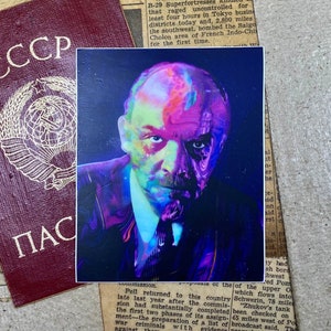 Könnte beinhalten: Eine Nahaufnahme eines alten Passes mit dem Wort "CCP" in roten Buchstaben auf dem Umschlag. Der Pass ist teilweise verdeckt von einem farbenfrohen Porträt eines Mannes mit einem ernsten Ausdruck. Das Porträt ist in einem lebendigen, abstrakten Stil mit Schattierungen von Pink, Blau und Grün gehalten.