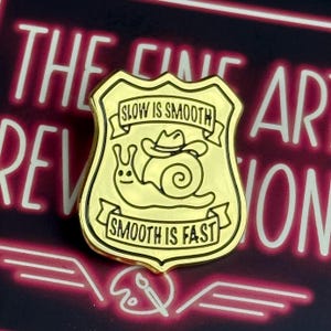 May include: A gold-colored enamel pin in the shape of a badge. The pin features a cartoon snail wearing a cowboy hat. The text "SLOW IS SMOOTH" and "SMOOTH IS FAST" are displayed on banners above and below the snail.