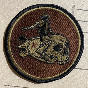 May include: A brown and green embroidered patch featuring a cartoon cowboy riding a skull with wings. The patch is circular and has a black border.