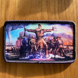 Op de afbeelding: Een kleurrijk geborduurd patch met een historische scène met een man die een geweer en een vlag vasthoudt. De scène speelt zich af tegen een achtergrond van een stormachtige lucht en een slagveld. De patch is omgeven door een zwarte rand.