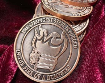 May include: A bronze-colored coin with the text "The Strongest Steel Is Forged In The Fire Of A Dumpster" around the edge. The center of the coin features a stylized image of a dumpster fire.