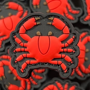 Puede incluir: Un parche de cangrejo rojo con costuras negras. El cangrejo está bordeado de negro y tiene un fondo negro.