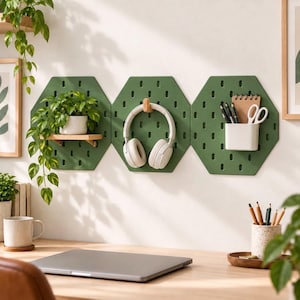 May include: Three olive green hexagonal pegboards mounted on a wall. The left board holds a small potted plant on a wooden shelf. The center board features a wooden hook holding white headphones. The right board has a white cup with pens and scissors.