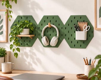 May include: Three olive green hexagonal pegboards mounted on a wall. The left board holds a small potted plant on a wooden shelf. The center board features a wooden hook holding white headphones. The right board has a white cup with pens and scissors.