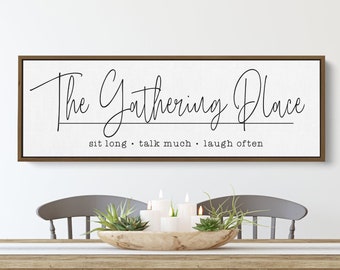 May include: A brown wooden framed sign with white background and black text that reads "The Gathering Place" with the tagline "sit long. talk much. laugh often". The sign is hanging on a white wall above a wooden table with gray chairs.