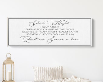 May include: A white wooden bench with a cream throw blanket and a decorative pillow. Two gold lanterns are on either side of the bench. A Christmas tree is on the left and another Christmas tree is on the right. A black and white sign with the text "Silent Night Holy Night Shepherds Quake at the Sight Glories Stream from Heaven Afar Heavenly Hosts Sign Alleluia Christ our Savior is born" is hanging on the wall behind the bench.