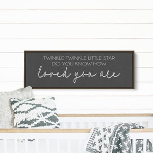 May include: A dark gray framed sign with white text that reads "Twinkle Twinkle Little Star Do You Know How Loved You Are". The sign is hanging on a white wood wall above a white crib with a gray and white blanket.