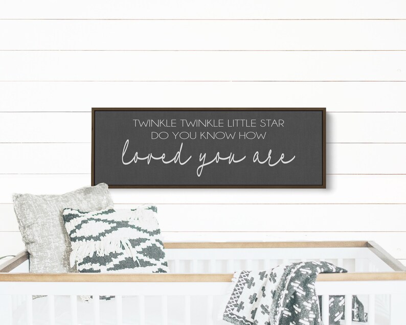 May include: A dark gray framed sign with white text that reads "Twinkle Twinkle Little Star Do You Know How Loved You Are". The sign is hanging on a white wood wall above a white crib with a gray and white blanket.