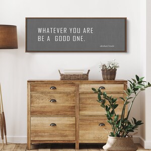 May include: A gray canvas sign with a wooden frame, featuring the quote "Whatever you are, be a good one." attributed to Abraham Lincoln.