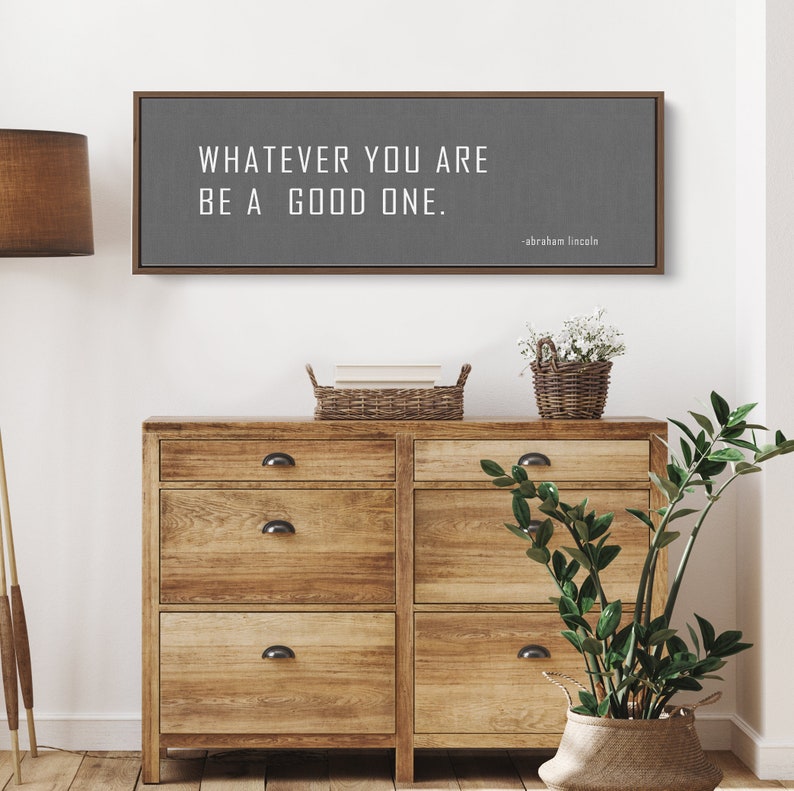 May include: A gray canvas sign with a wooden frame, featuring the quote "Whatever you are, be a good one." attributed to Abraham Lincoln.