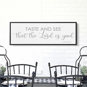 May include: A black framed canvas wall art with the text "Taste and see that the Lord is good" and the scripture reference "Psalm 34:8". The canvas is hanging on a white brick wall above a black dining table with black chairs and a black and white checkered table runner.