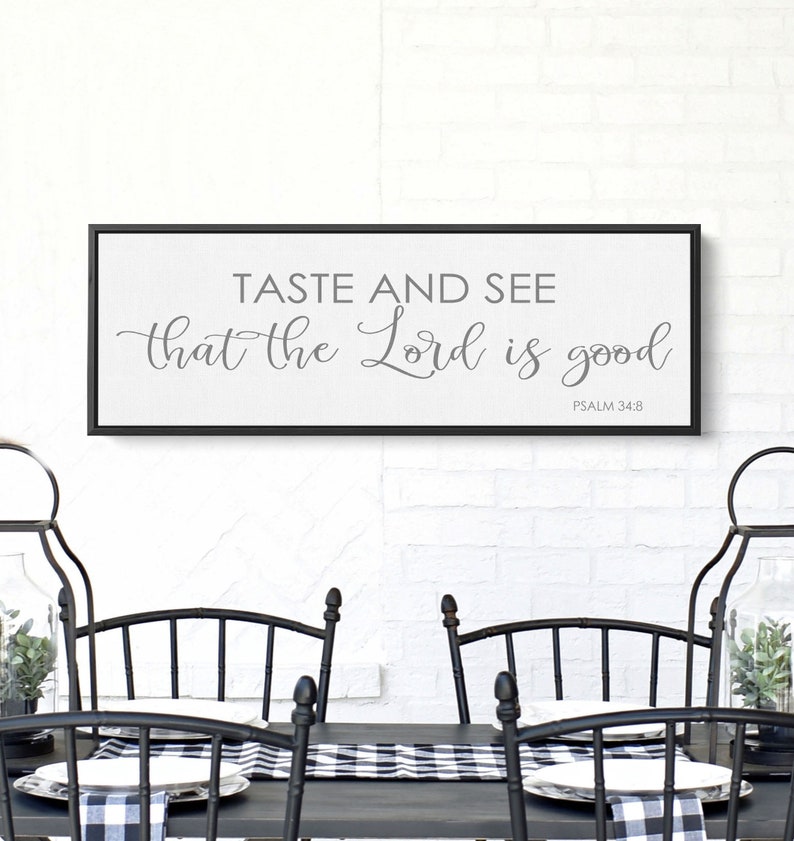 May include: A black framed canvas wall art with the text "Taste and see that the Lord is good" and the scripture reference "Psalm 34:8". The canvas is hanging on a white brick wall above a black dining table with black chairs and a black and white checkered table runner.