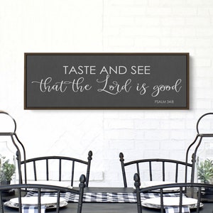 May include: A framed sign with a gray background and a white inscription that reads "Taste and see that the Lord is good, Psalm 34:8". The sign is hanging on a white brick wall above a black table with black chairs.