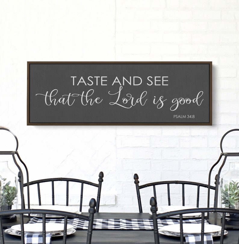 May include: A framed sign with a gray background and a white inscription that reads "Taste and see that the Lord is good, Psalm 34:8". The sign is hanging on a white brick wall above a black table with black chairs.
