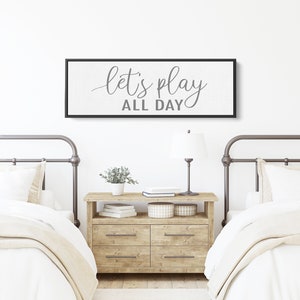 May include: A black framed canvas sign with white background and gray text that reads "let's play all day" hanging above two twin beds with white bedding and metal headboards.