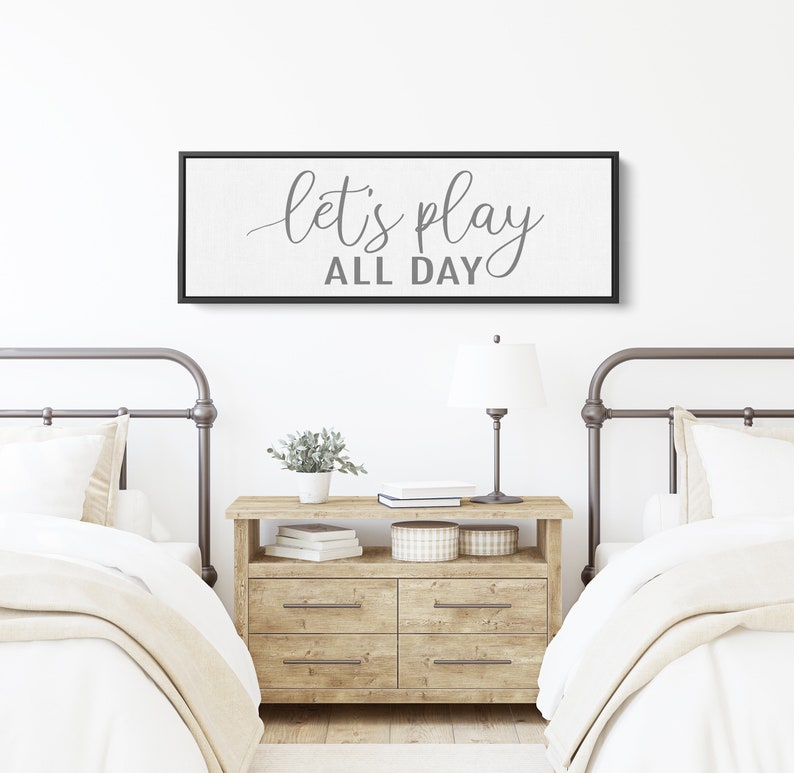 May include: A black framed canvas sign with white background and gray text that reads "let's play all day" hanging above two twin beds with white bedding and metal headboards.