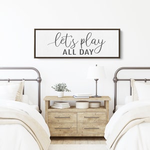 May include: A white canvas sign with a brown wood frame hangs above two twin beds. The sign reads "let's play all day" in a gray cursive font.