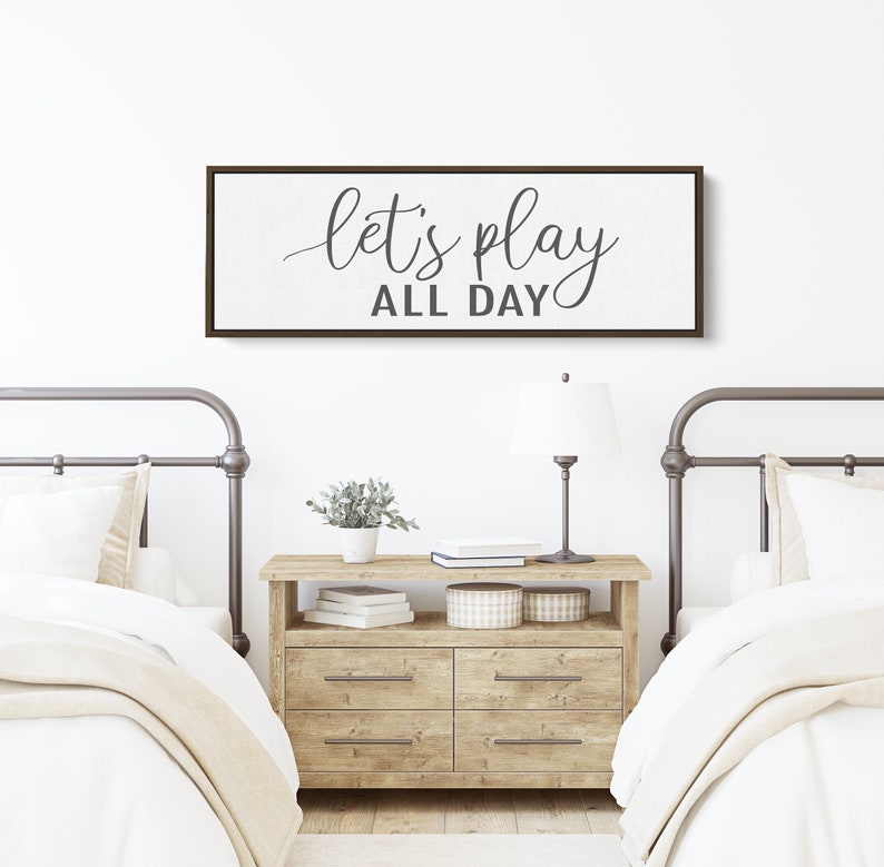 May include: A white canvas sign with a brown wood frame hangs above two twin beds. The sign reads "let's play all day" in a gray cursive font.