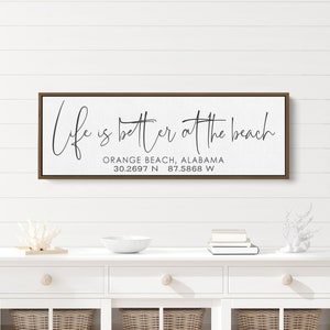 May include: A brown wooden sign with white canvas and black text that reads "Life is better at the beach" with the location "Orange Beach, Alabama 30.2697 N 87.5868 W" underneath.