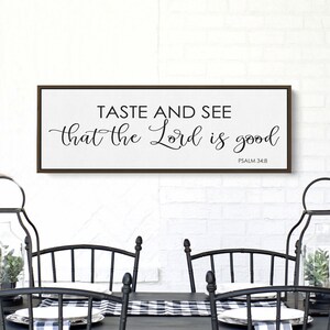 May include: A framed wood sign with the text "Taste and see that the Lord is good" and the scripture reference "Psalm 34:8". The sign is hanging on a white brick wall above a black table with two black chairs.