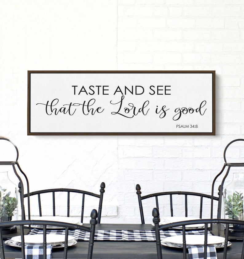 May include: A framed wood sign with the text "Taste and see that the Lord is good" and the scripture reference "Psalm 34:8". The sign is hanging on a white brick wall above a black table with two black chairs.