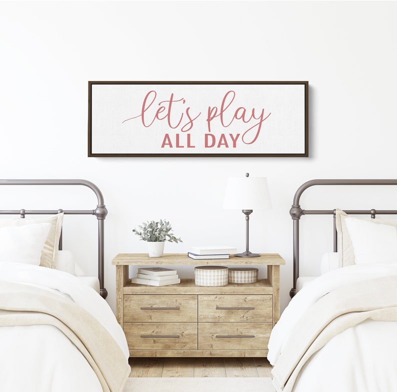 May include: A wooden sign with the text "Let's play all day" in pink script on a white background. The sign is hanging on a white wall above a wooden dresser with two beds on either side.