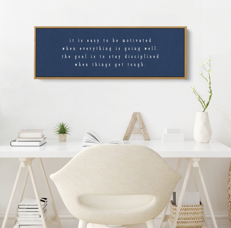 K&ouml;nnte beinhalten: Ein gerahmter Leinwanddruck in Marineblau mit wei&szlig;em Text, der lautet "it is easy to be motivated when everything is going well. the goal is to stay disciplined when things get tough."