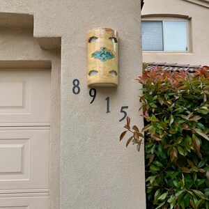 May include: A cylindrical, yellow ceramic address light fixture with a turquoise and brown geometric design. The fixture is mounted on a beige stucco wall, with the numbers "8915" below it. The fixture has several cut-out openings.