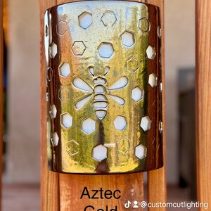 May include: Gold-colored metal wall sconce with a honeycomb pattern and a bee design. The sconce is mounted on a wooden surface. Aztec Gold. @customcutlighting