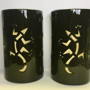 May include: Two cylindrical dark olive green ceramic candle holders featuring a cutout design of a lizard on the front. The glossy surface of the vessels contrasts with the light-colored interior visible through the lizard-shaped openings.