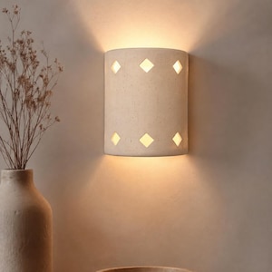 May include: A semi-cylindrical wall sconce made of off-white speckled material featuring six diamond-shaped cutouts arranged in two horizontal rows. The fixture emits a warm, soft glow from the top, bottom, and through the geometric openings.