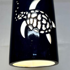 May include: A black pendant light with a sea turtle design. The turtle is cut out of the black shade, allowing light to shine through. The light fixture hangs from a silver chain and has a warm, yellow glow.