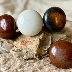 May include: Four polished, spherical stones in shades of brown, white, and black rest on a textured, light-brown surface. The stones vary in color and sheen, with some appearing glossy and others more matte.