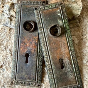 May include: Two antique bronze door plates with keyholes and circular knob openings. The rectangular plates have ornate, raised borders and show signs of age and wear, with a mix of bronze and green patina. The plates are likely from a vintage door.