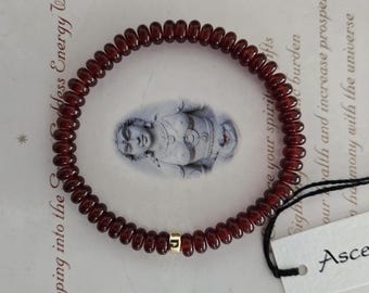 May include: A beaded bracelet made of small, round, dark red gemstones. A small gold-colored bead is centered on the bracelet. The bracelet is on a patterned background with text and a tag that says "Ascen".