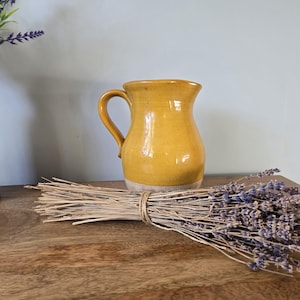 May include: A handmade yellow ceramic pitcher with a handle and a light-colored base. A bundle of dried stems and lavender flowers are on a wooden surface. The pitcher is the focal point, showcasing its unique shape and color.