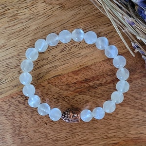 May include: White gemstone beaded bracelet with a copper Buddha head charm. The bracelet is made of smooth, round beads and is strung on a clear elastic cord.