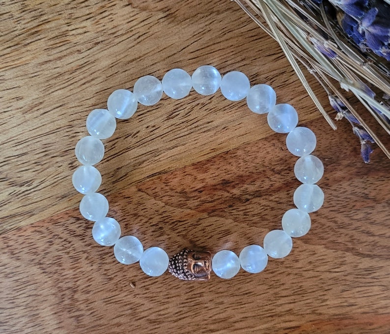 May include: White gemstone beaded bracelet with a copper Buddha head charm. The bracelet is made of smooth, round beads and is strung on a clear elastic cord.