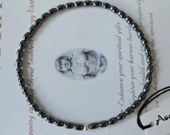 May include: A circular beaded necklace composed of small, polished, dark grey metallic oval beads arranged in a continuous loop, featuring a single small, round, silver-toned bead at the bottom centre, attached to a white tag with black text.