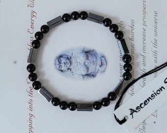 May include: Black beaded bracelet with rectangular hematite accents. The bracelet is on a white surface with text that reads "Tapping into the Less Energy Within". A tag reads "Ascension S".