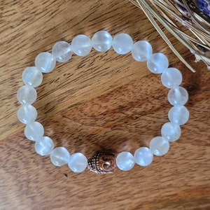 May include: White gemstone bracelet with a copper Buddha charm. The bracelet is made of round beads and is strung on elastic.