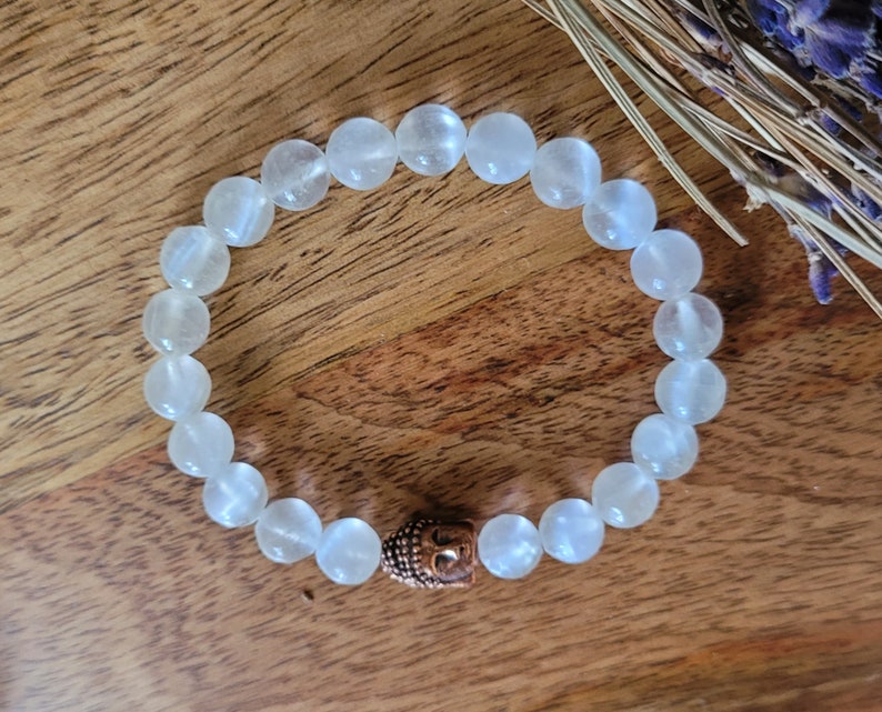 May include: White gemstone bracelet with a copper Buddha charm. The bracelet is made of round beads and is strung on elastic.