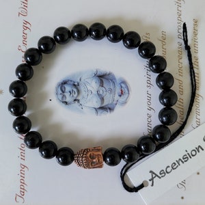 May include: Black beaded bracelet with a copper Buddha head charm. The bracelet is tied with a black cord and has a tag that reads "Ascension Stone".