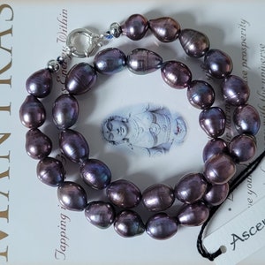 Op de afbeelding: Een ketting van donkerpaarse zoetwaterparels met een zilveren sluiting. De parels zijn iriserend en hebben een licht ovale vorm. De ketting ligt op een boek met het woord "MANTRAS" op de kaft.