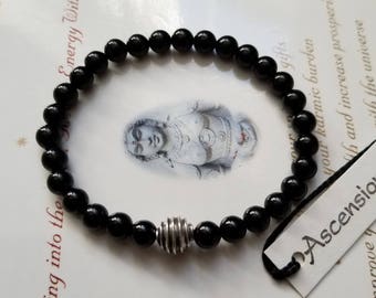 May include: Black beaded bracelet with a silver metal charm. The bracelet is on a white surface with a white card that says "Ascension Stones" in black text.