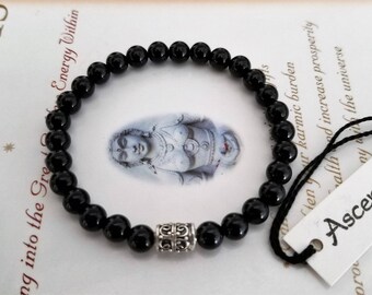 May include: Black beaded bracelet with a silver-colored spacer bead. The bracelet is displayed on a printed surface with text and an image of a figure. A tag with the word "Ascensi" is attached.