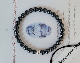 Puede incluir: Un brazalete de cuentas de hematites negras con un dije de plata. El brazalete está sobre un fondo blanco con texto que dice "Tapping into the Goddess Energy With" y "Ascension Shop".