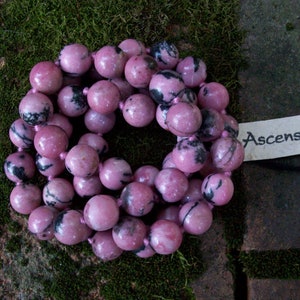 May include: A strand of pink and black gemstone beads. The beads are round and have a smooth, polished finish. The beads are strung on a clear string. The text "Ascension Stone" is visible on a white tag.